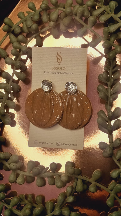 Pair of gold earrings on a brand card with green foliage in the background