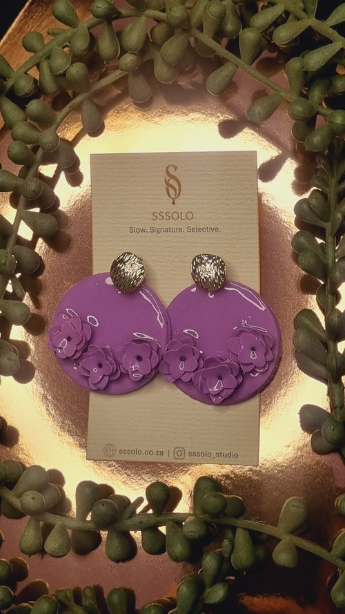 Purple earrings on a card with gold studs, leafy decorated background 