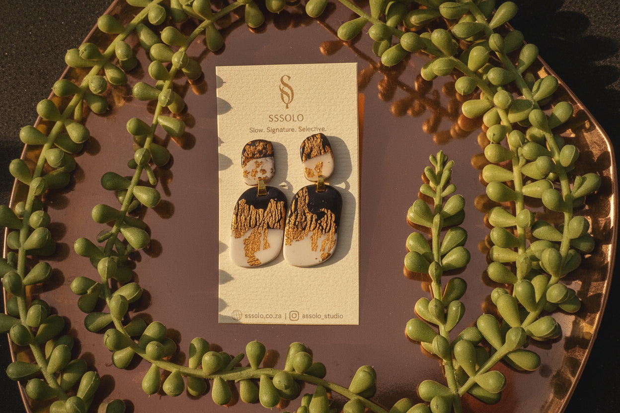 Gold and black earrings on a branded card with green leaves around