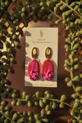 Pair of pink earrings with gold accents on a card, surrounded by green leaves.