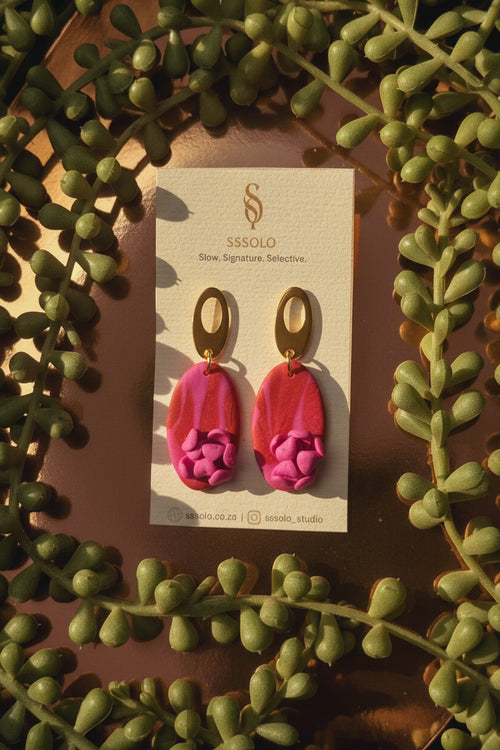 Pair of pink earrings with gold accents on a card, surrounded by green leaves.