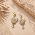 A pair of gold-tone dangle earrings with irregular white stone-like charms filled with gold leaf and a small clear crystal drop, styled on a beige surface with a textured stone slab and dried palm leaf.