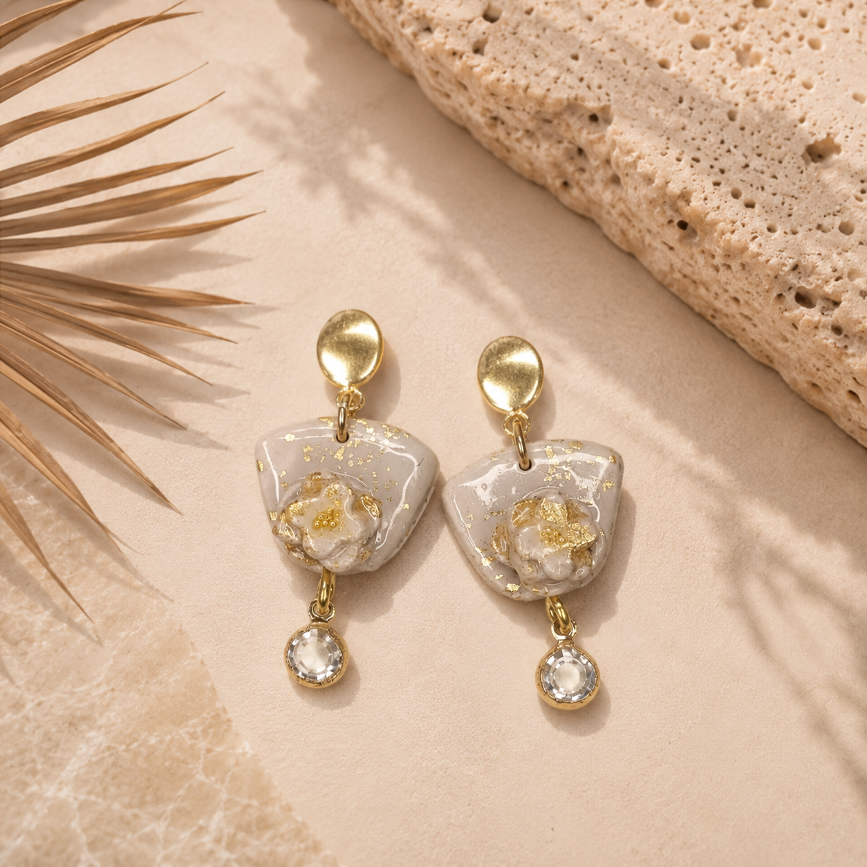 A pair of gold-tone dangle earrings with irregular white stone-like charms filled with gold leaf and a small clear crystal drop, styled on a beige surface with a textured stone slab and dried palm leaf.